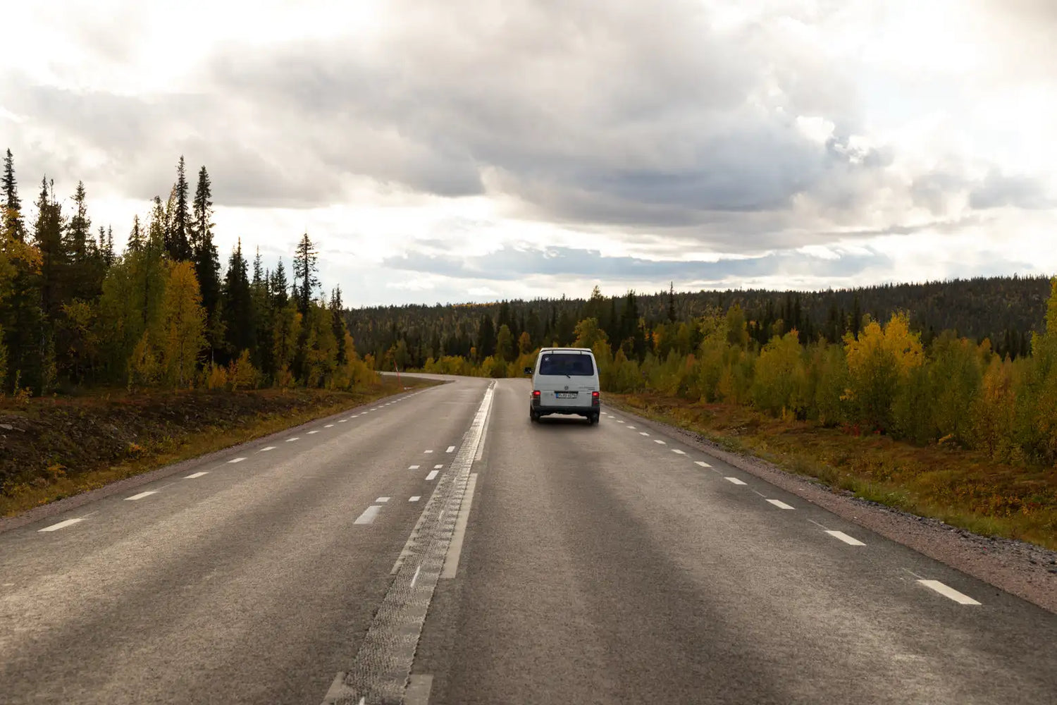 Ein weißer Transporter fährt eine kurvenreiche Straße entlang, gesäumt von Bäumen in Herbstfarben unter einem wolkenverhangenen Himmel.